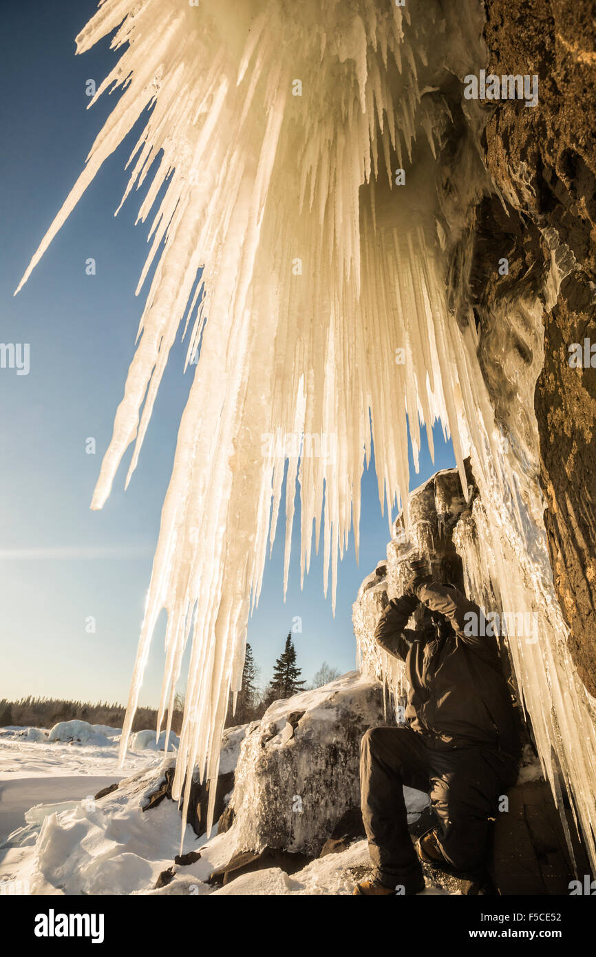 Lato basso angolo POV gigante di ghiaccioli appesi da una scogliera rocciosa con una persona in abbigliamento invernale inginocchiato sotto ghiaccioli Foto Stock
