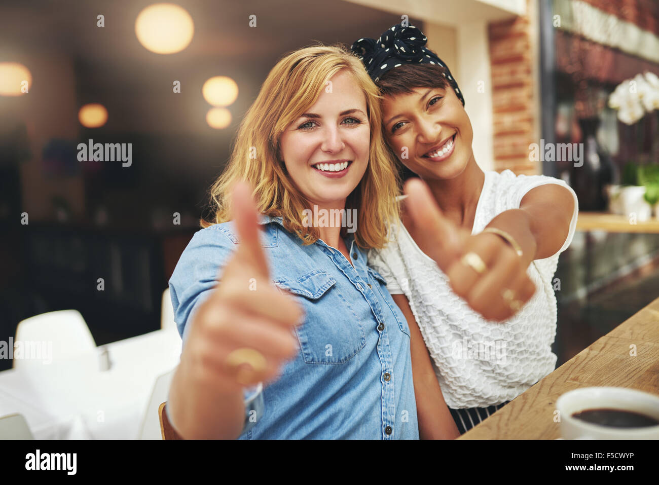 Due giovani felici amici femminili dando un pollice in alto gesto di approvazione e di successo come essi sedersi sotto braccio in una caffetteria godendo Foto Stock