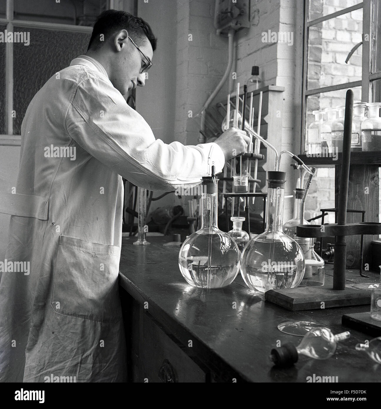 Degli anni Cinquanta, storico, maschio scienziato in camice bianco a lavorare in un laboratorio di chimica, England, Regno Unito, la miscelazione di liquidi differenti. Foto Stock