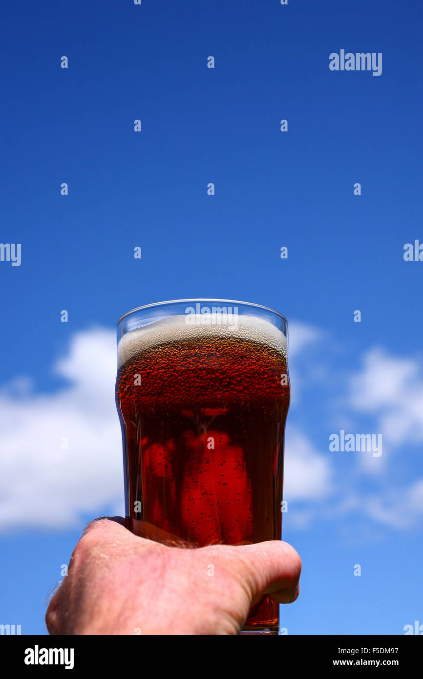 Man mano che tiene una pinta bicchiere di birra amara contro un Cielo di estate blu Foto Stock
