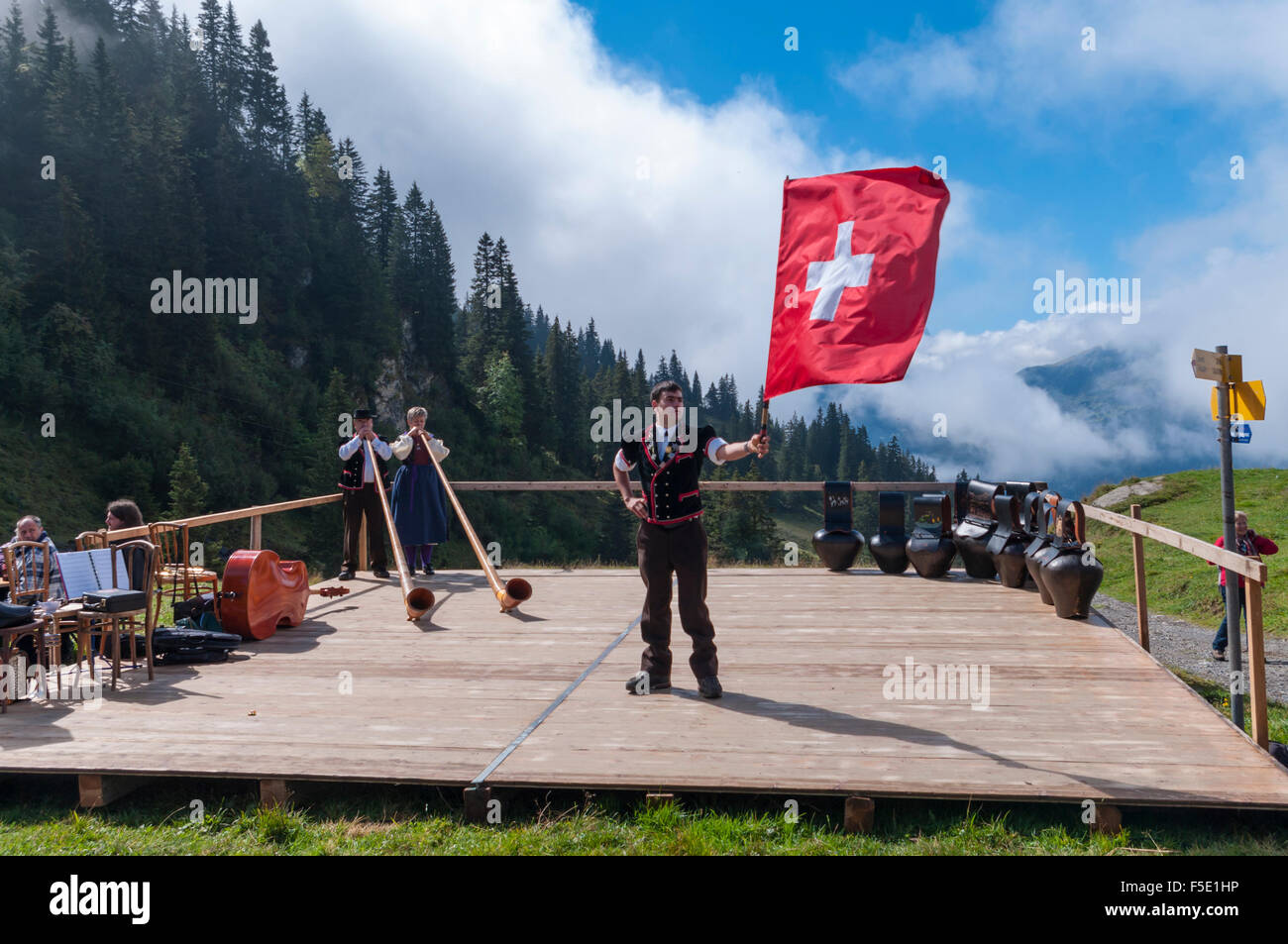 Giovane uomo in Oberland costumi folk dando un flag di gettare le prestazioni nelle Alpi Svizzere. In background alphorns e campane delle mucche. Foto Stock