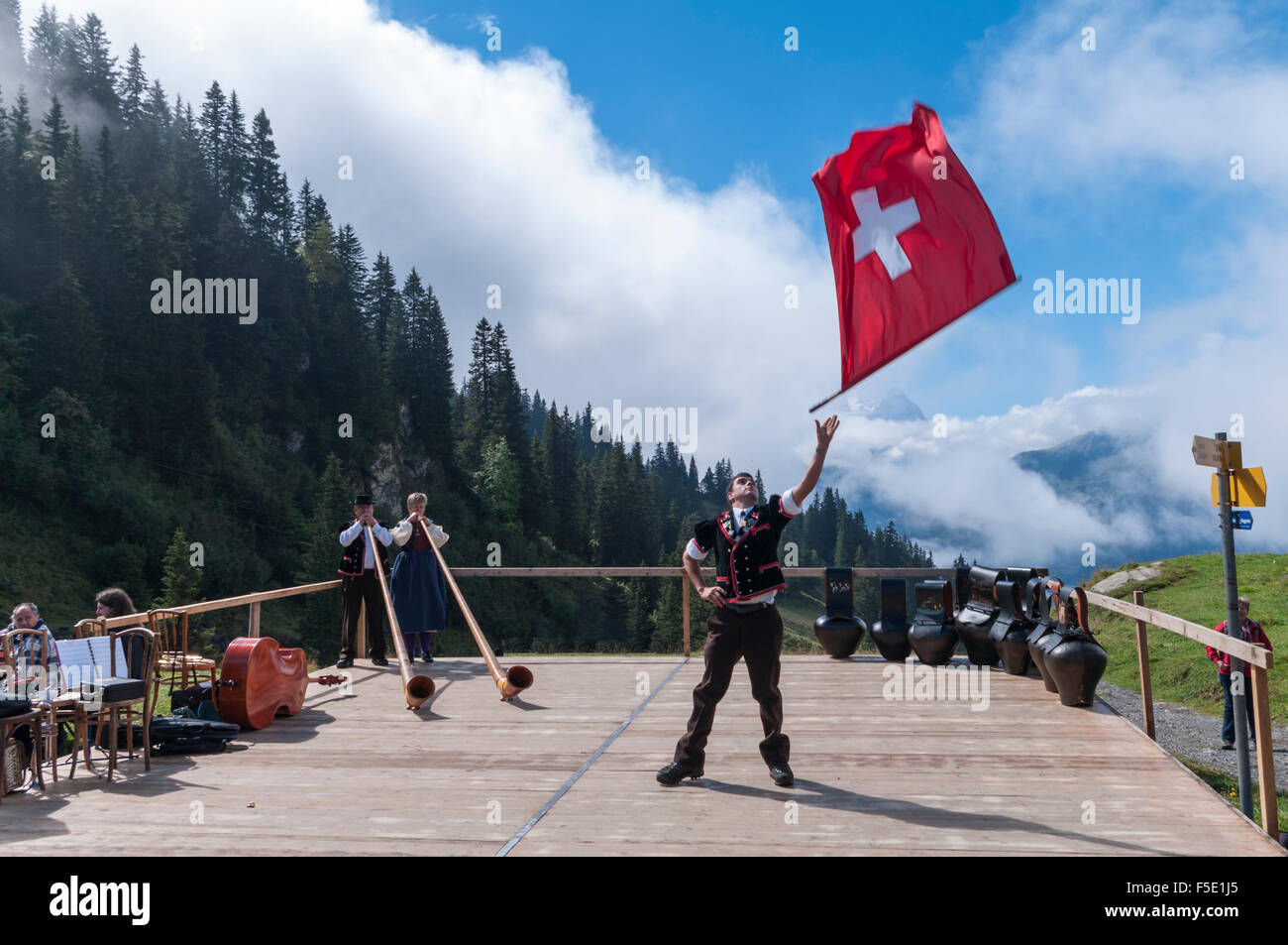 Giovane uomo in oberland costumi folk dando un flag di gettare le prestazioni. In background alphorns e campane delle mucche. Foto Stock