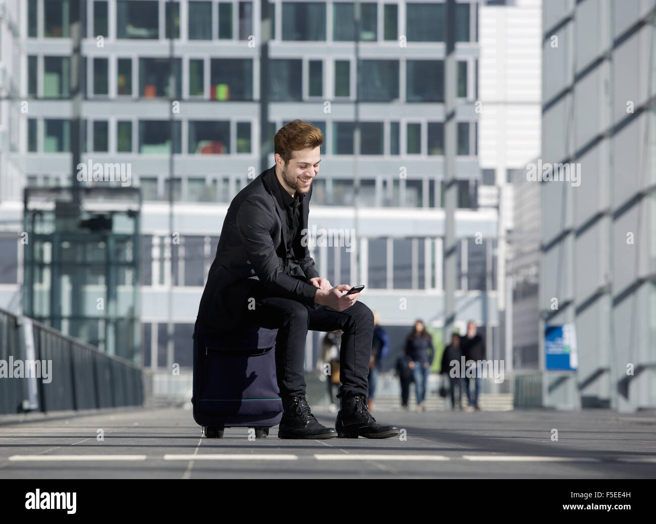 Ritratto di un allegro giovane uomo seduto sulla valigia alla stazione guardando al telefono cellulare Foto Stock