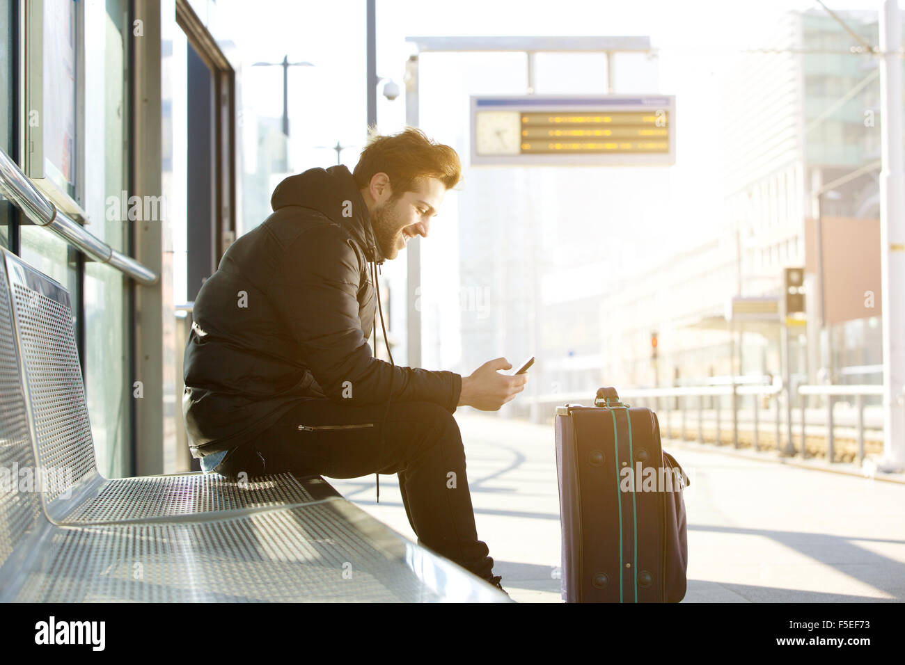 Ritratto laterale di un sorridente giovane uomo seduto con il telefono cellulare ed il sacco in attesa del treno Foto Stock
