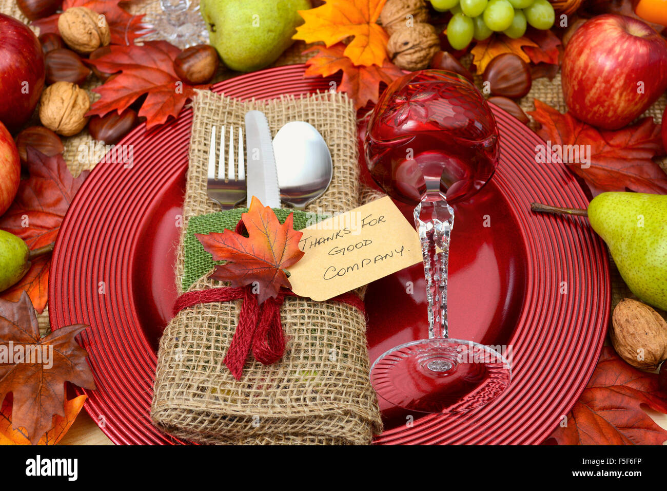Ringraziamento rustico con tela avvolta posate, cristallo rosso bicchiere di vino sulla grande piastra caricabatterie e raccolto autunnale frutta e noci. Foto Stock