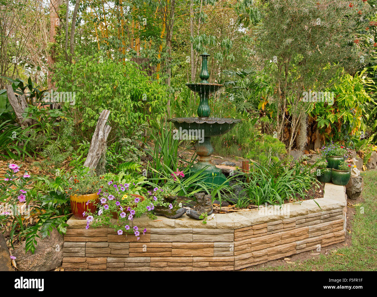 Giardino decorativo caratteristica con un basso muro di pietra, fontana ornata, piante in contenitori e orlate da prato, fiori, arbusti, alberi Foto Stock