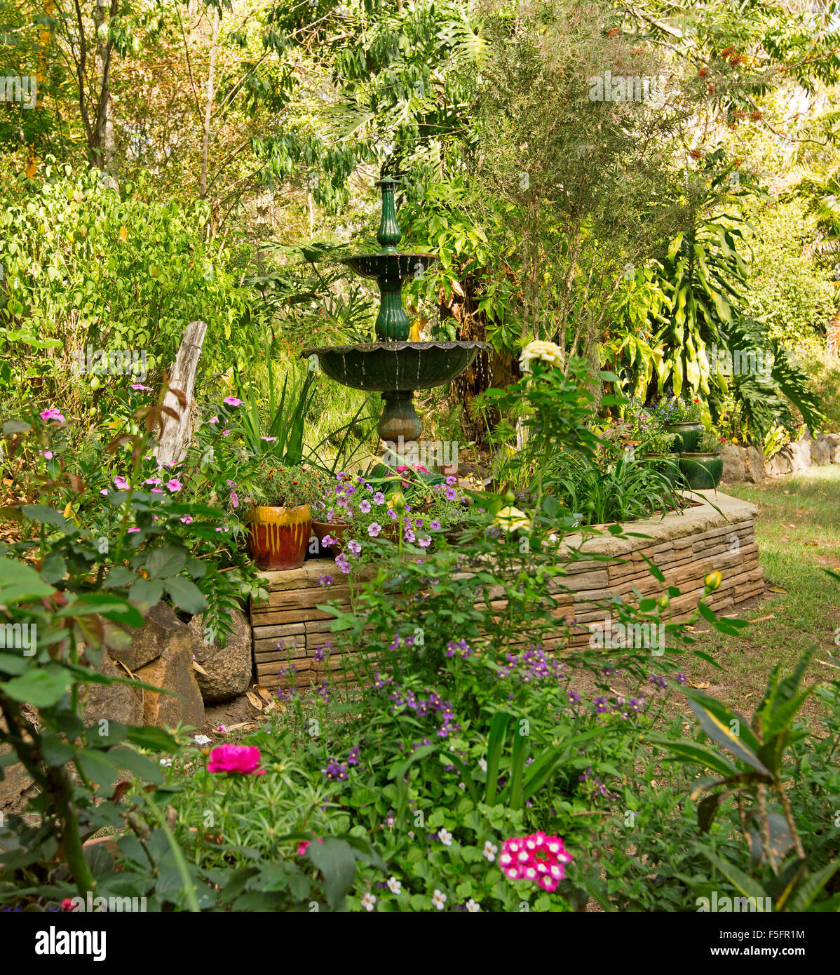 Giardino decorativo caratteristica con un basso muro di pietra, fontana ornata, piante in contenitori e orlate da prato, fiori, arbusti, alberi Foto Stock