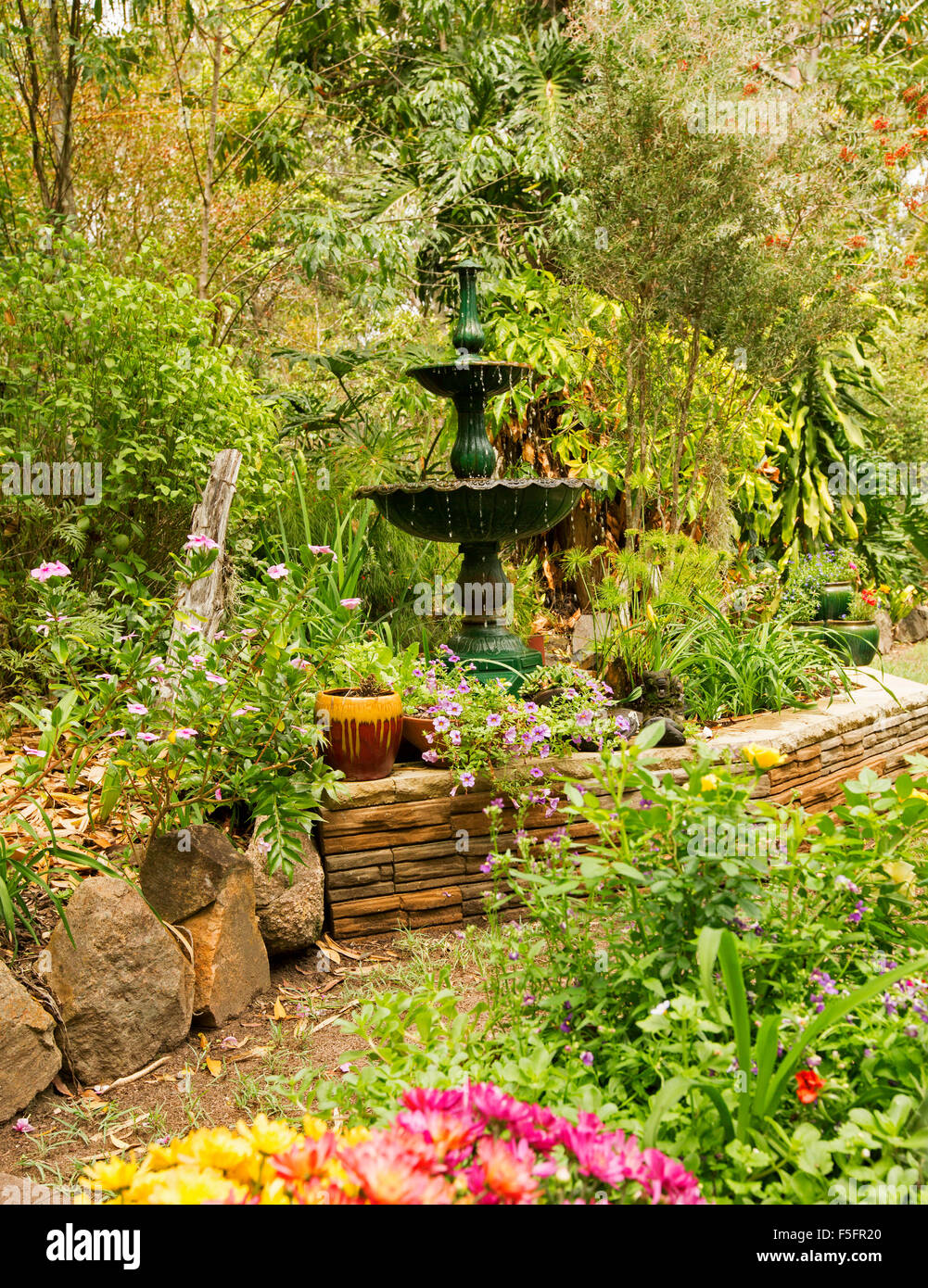 Giardino decorativo caratteristica con un basso muro di pietra, fontana ornata & piante in contenitori, orlata da arbusti, gli alpinisti e gli alti alberi Foto Stock