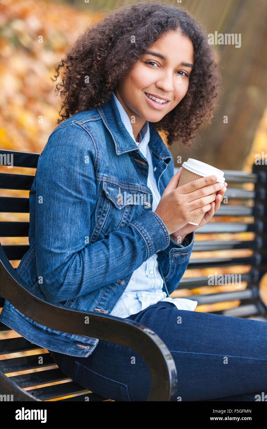 Bella felice razza mista americano africano ragazza adolescente femmina giovane donna a bere il caffè da asporto seduti su una panchina nel parco Foto Stock