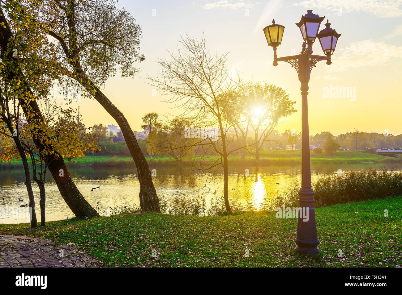 Paesaggio nel parco con alberi secolari e lampade vicino al lago al tramonto in autunno Foto Stock