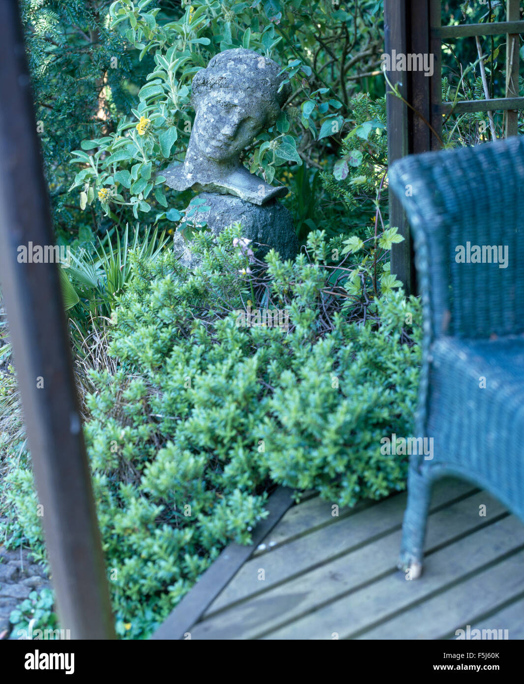 Dipinto di blu sedia in vimini su decking accanto a una testa in pietra sotto-coltivati con piante verdi in un giardino estivo Foto Stock