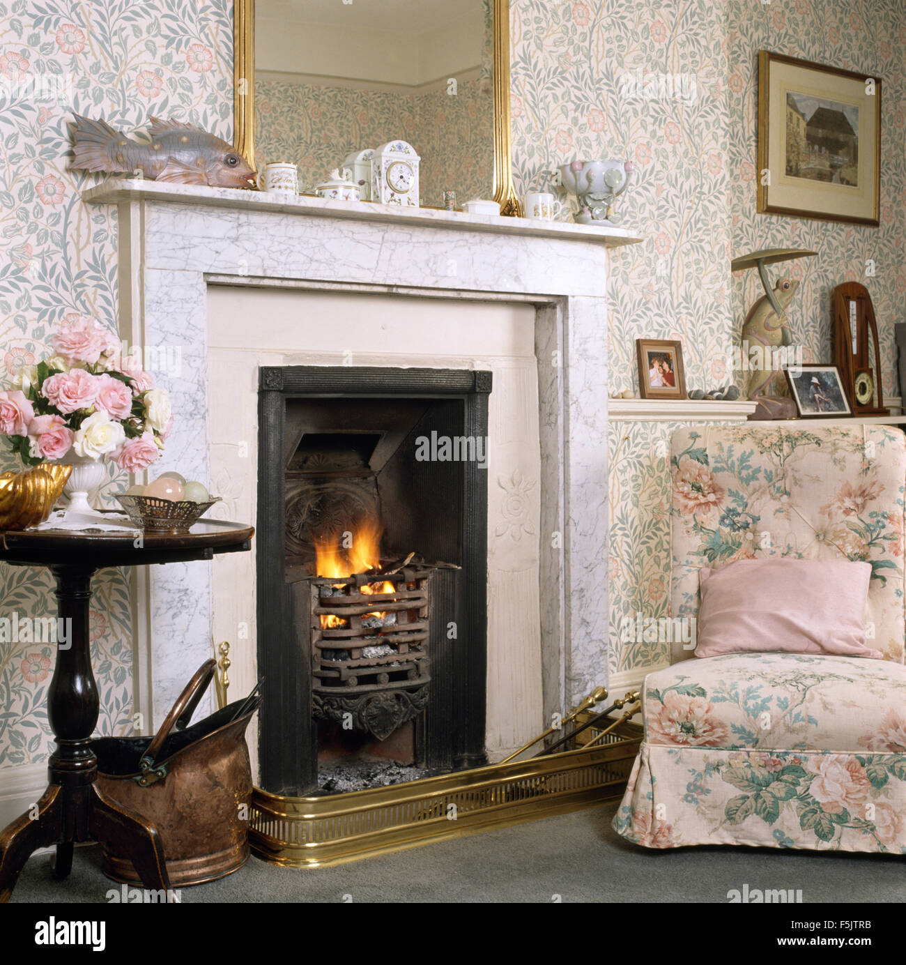 Sedia floreali accanto al caminetto con fuoco illuminato in salotto con carta da parati floreale Foto Stock
