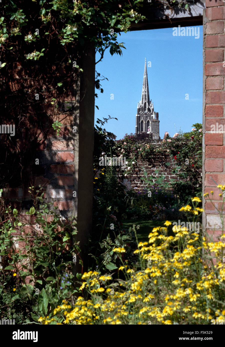 Veduta di una chiesa guglia attraverso una porta aperta in una parete del giardino con giallo heleniums crescente accanto alla porta Foto Stock