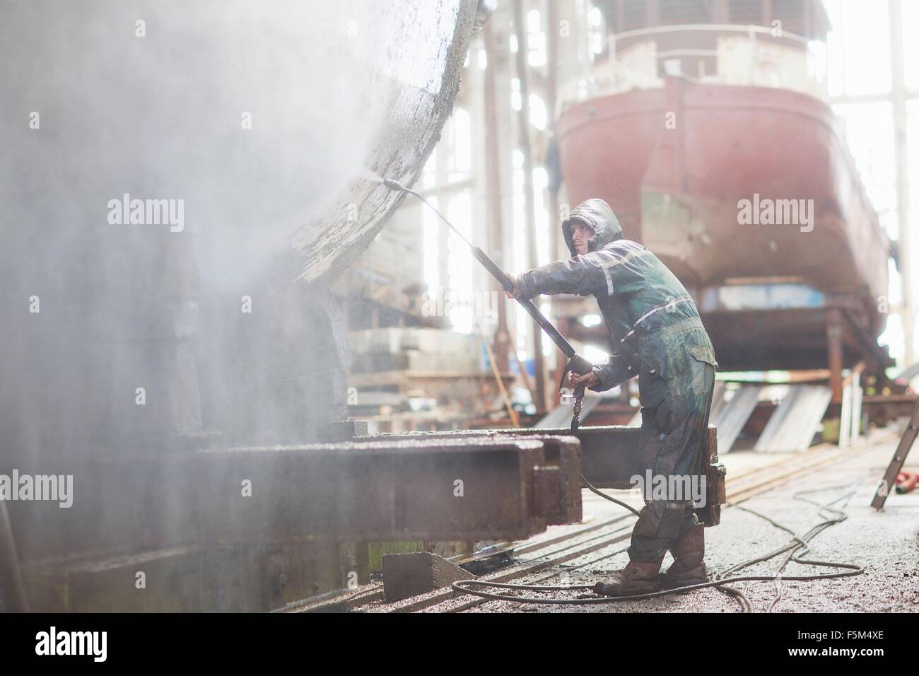 Lavoratore con tubo flessibile ad alta pressione in barca nel workshop di cantiere Foto Stock