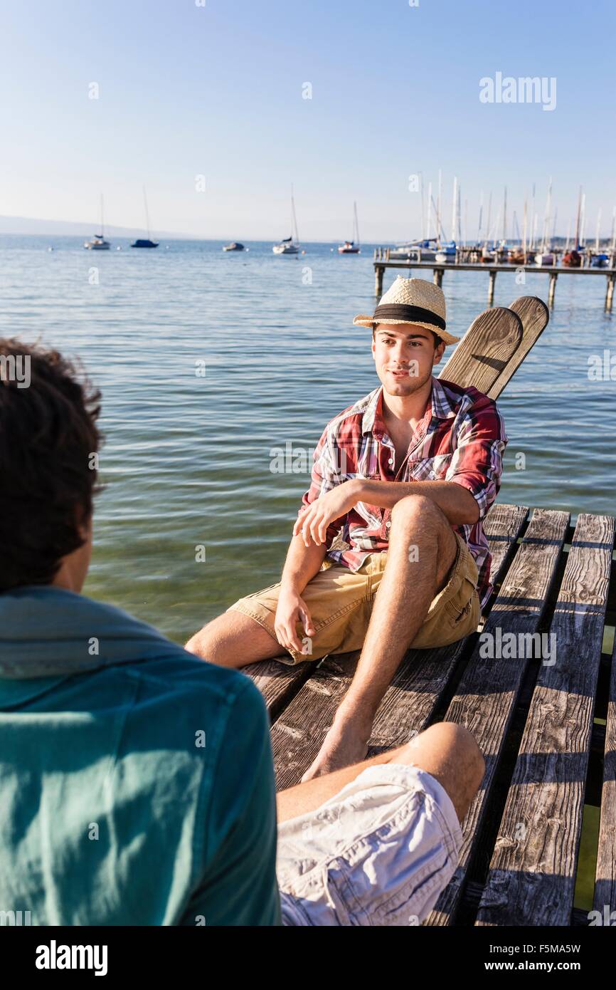 Amici seduti faccia a faccia sul molo di legno accanto al lago, Schondorf, Ammersee, Baviera, Germania Foto Stock