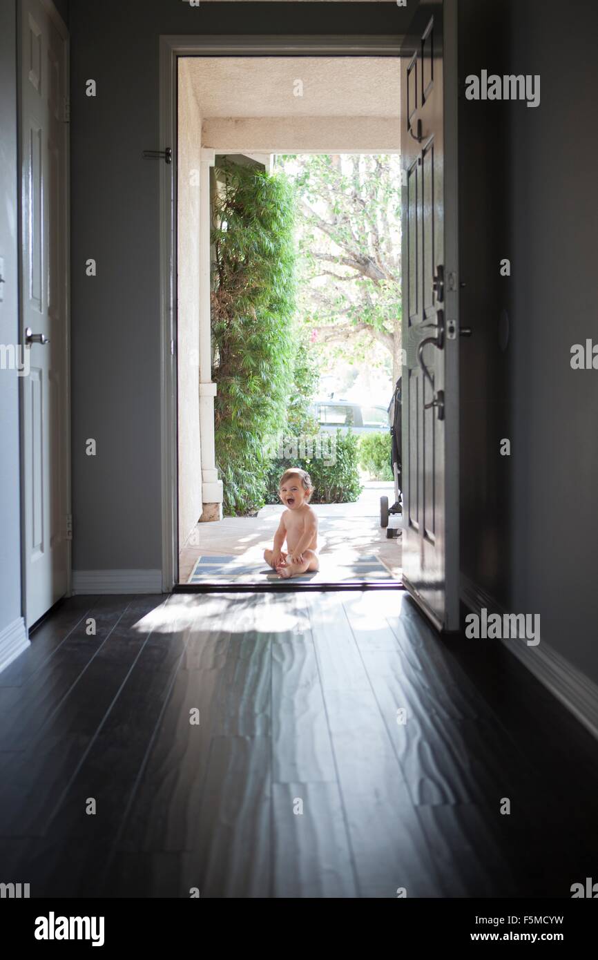 Visualizzazione attraverso l'ingresso del bambino seduto fuori ad aprire la  porta anteriore, guardando la telecamera con imboccatura aperta Foto stock  - Alamy