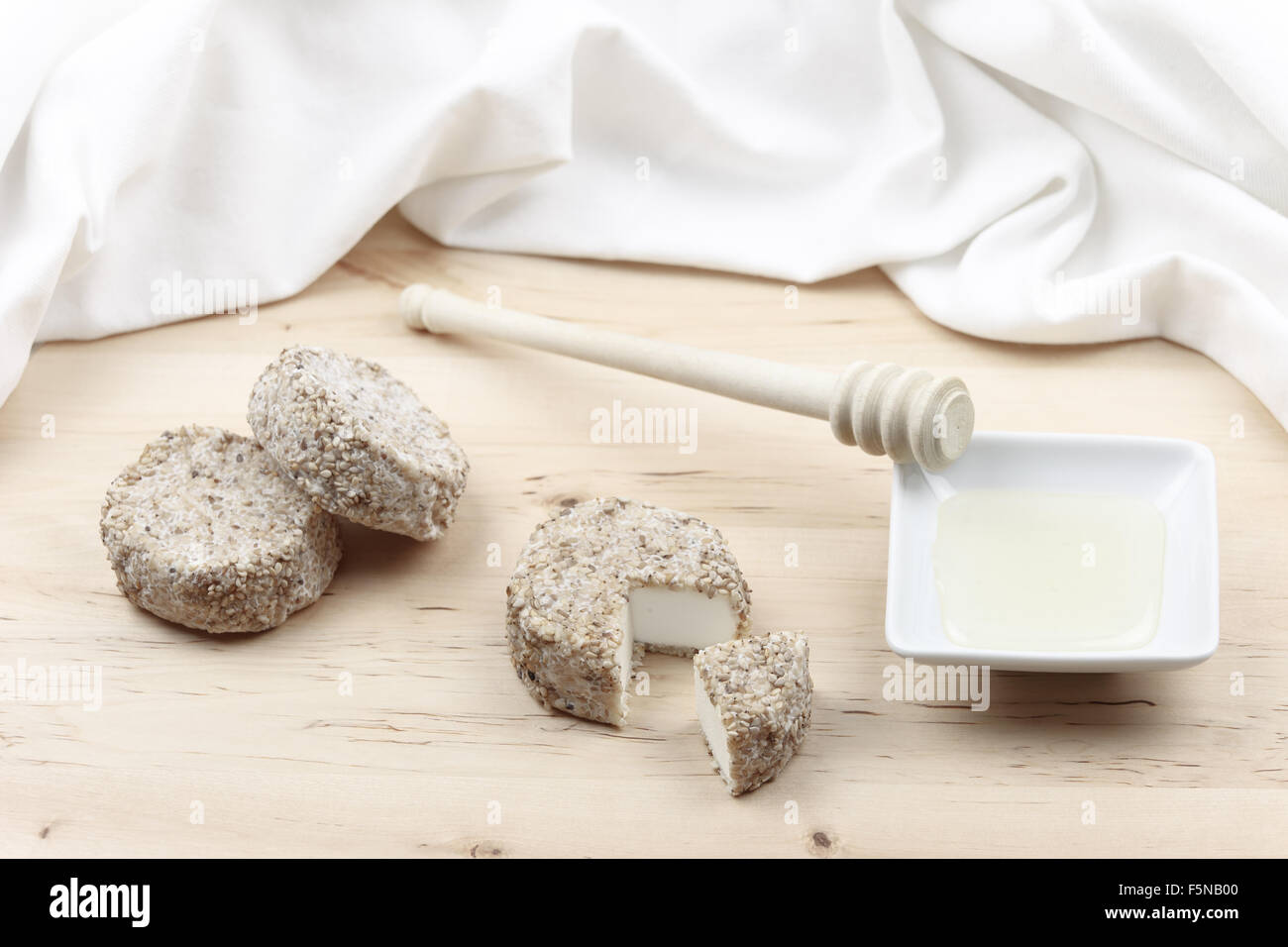 Formaggio di capra coperti con i semi di sesamo e miele e il suo bilanciere sul tagliere di legno e tovaglia bianca dello sfondo. Foto Stock