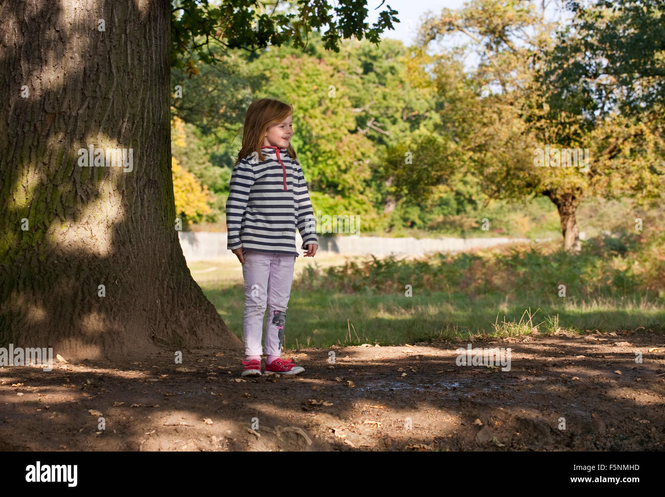 Una giovane ragazza sorge nell'ombra di un grande albero sorridente a qualcosa al di fuori dell'immagine. Essa è fiera con i capelli di zenzero. Foto Stock