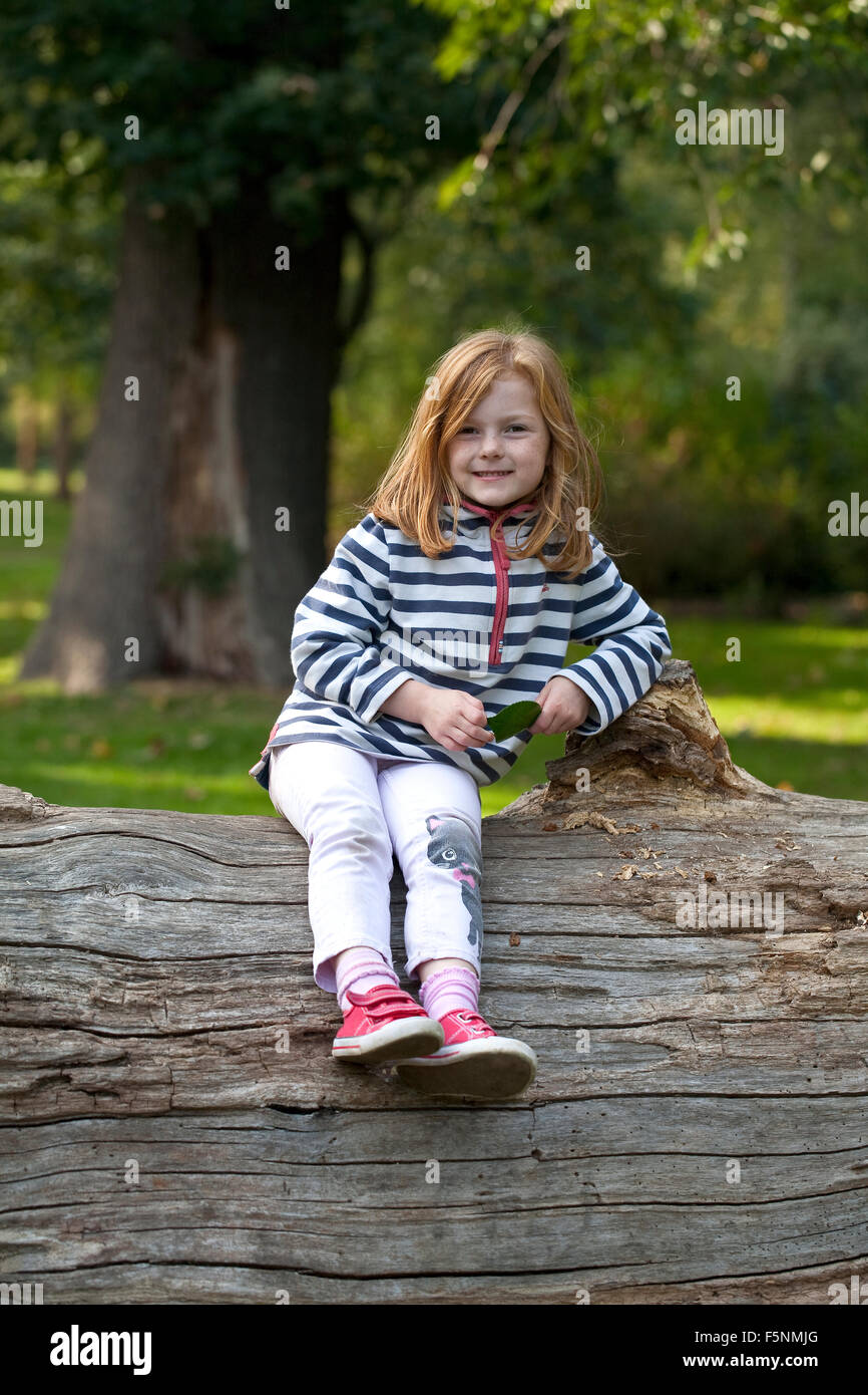 Una giovane ragazza con i capelli di zenzero è sorridente ampiamente come lei si siede in alto su un albero caduto trunk. Foto Stock