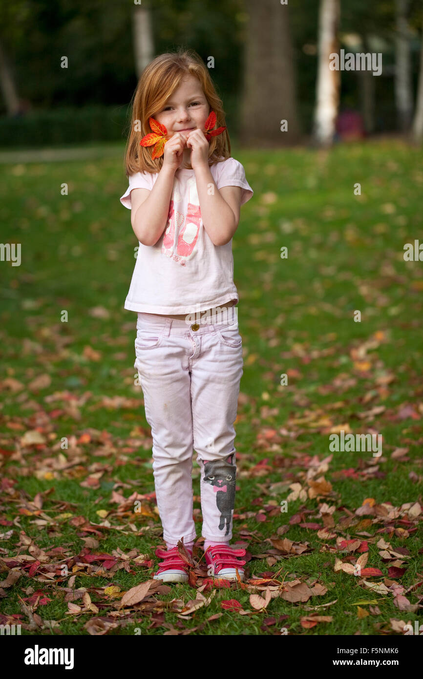 Una giovane ragazza con i capelli di zenzero contiene due belle foglie di autunno contro il suo volto come lei gioca su una soleggiata giornata autunnale. Foto Stock