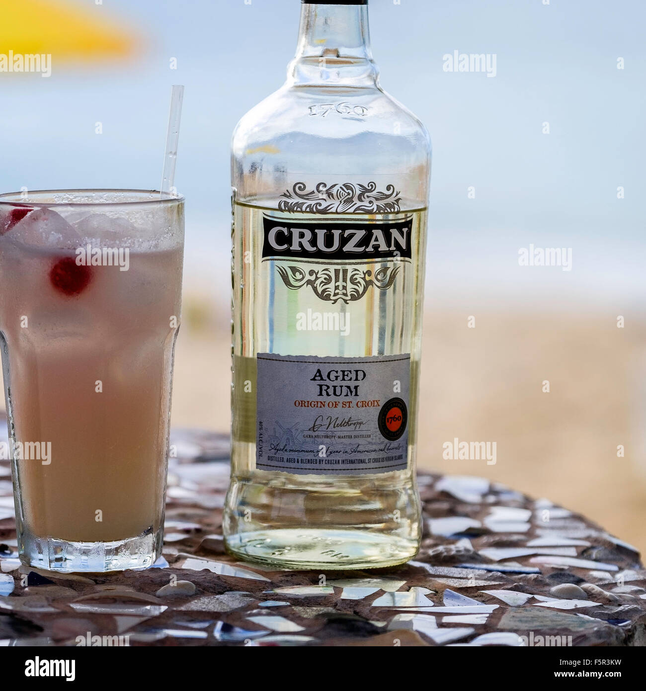 Una bottiglia di Cruzan Rum invecchiato e un cocktail di rum in un bicchiere seduti a un tavolo con un ombrellone da spiaggia e il mare dei Caraibi. Foto Stock