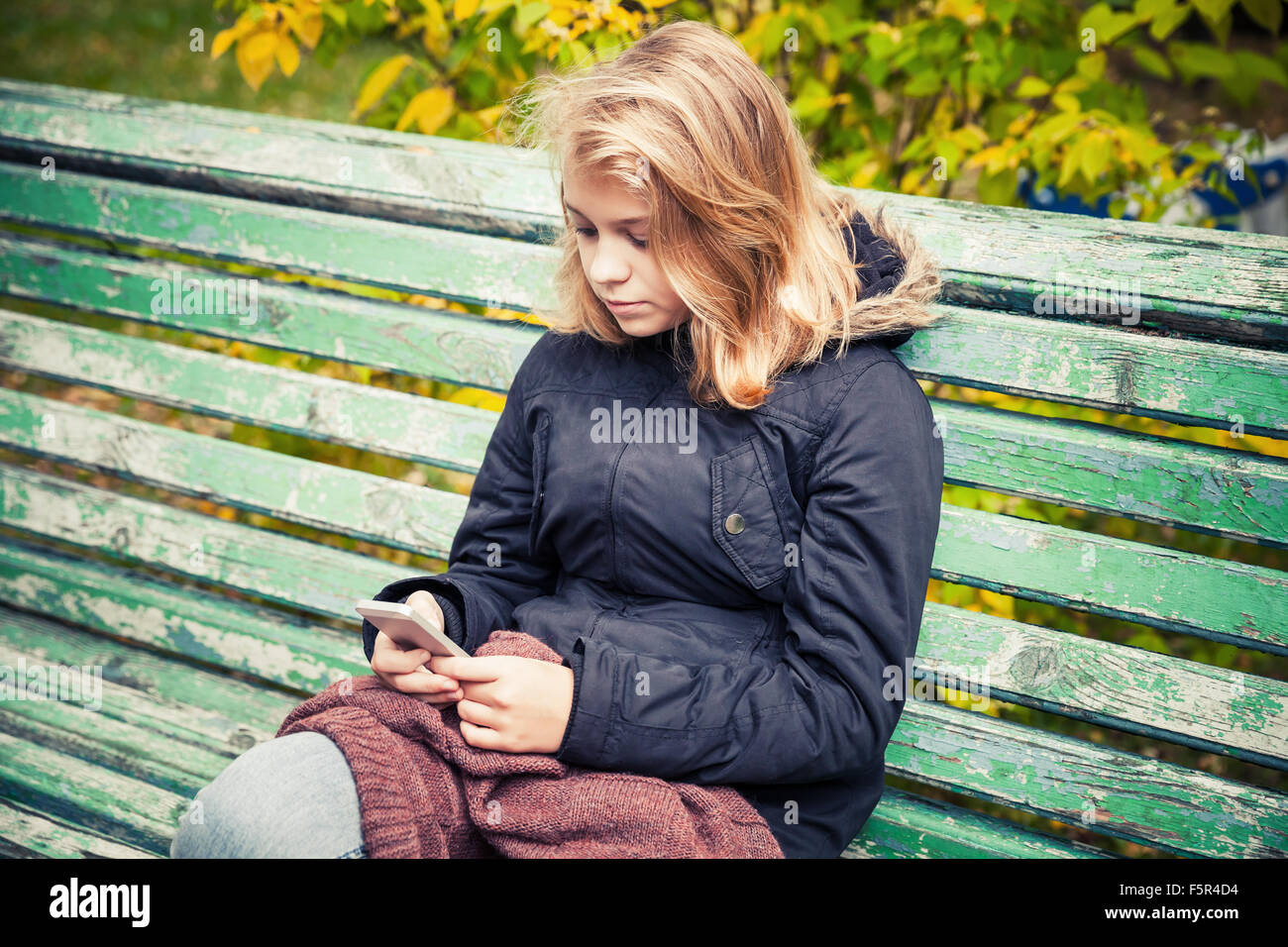 Bella bionda caucasica ragazza adolescente in giacca nera seduta sul vecchio parco verde di banco con smart phone in mani Foto Stock