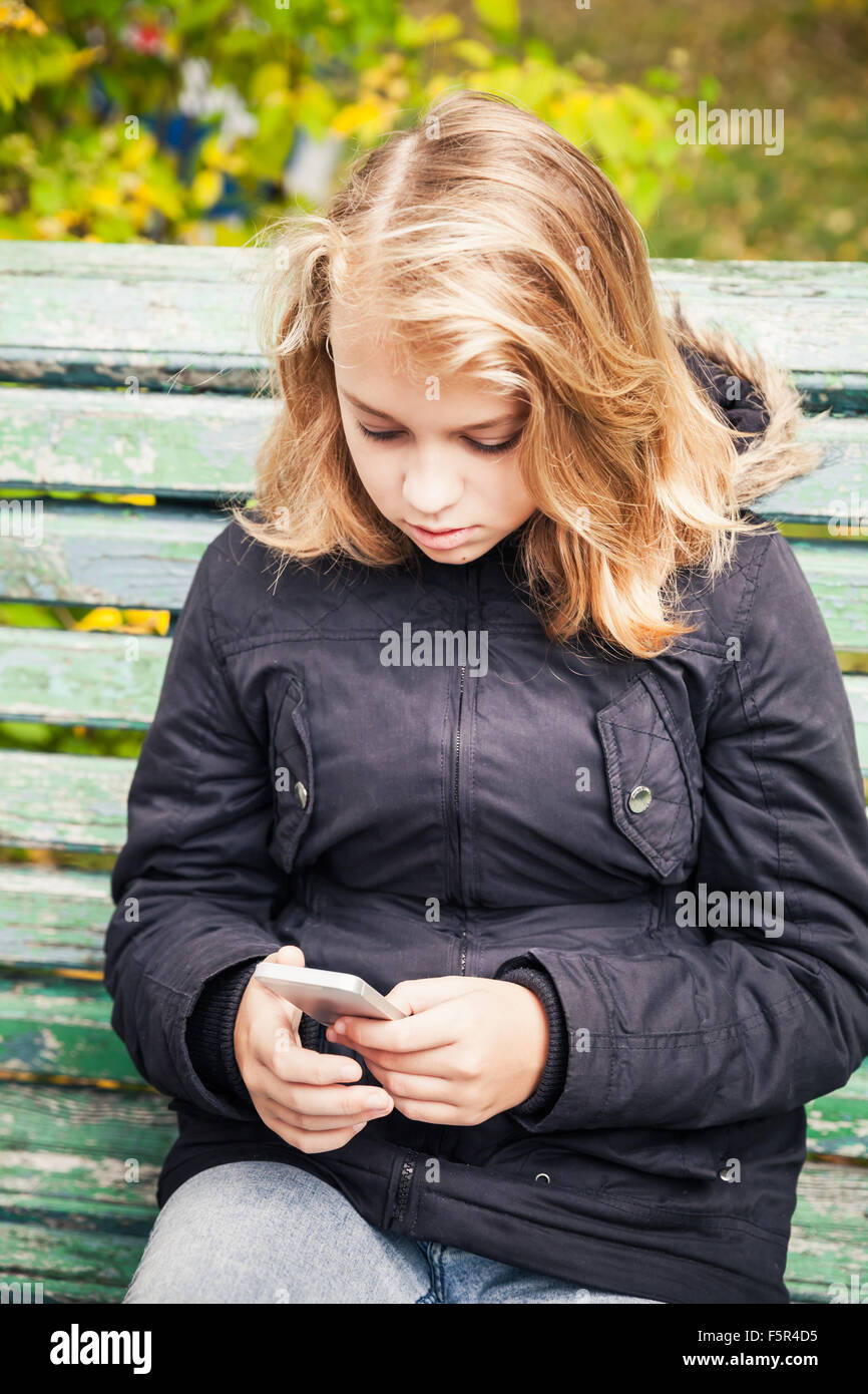 Bella bionda caucasica ragazza adolescente in giacca nera seduta sul banco di verde con lo smartphone in mani Foto Stock