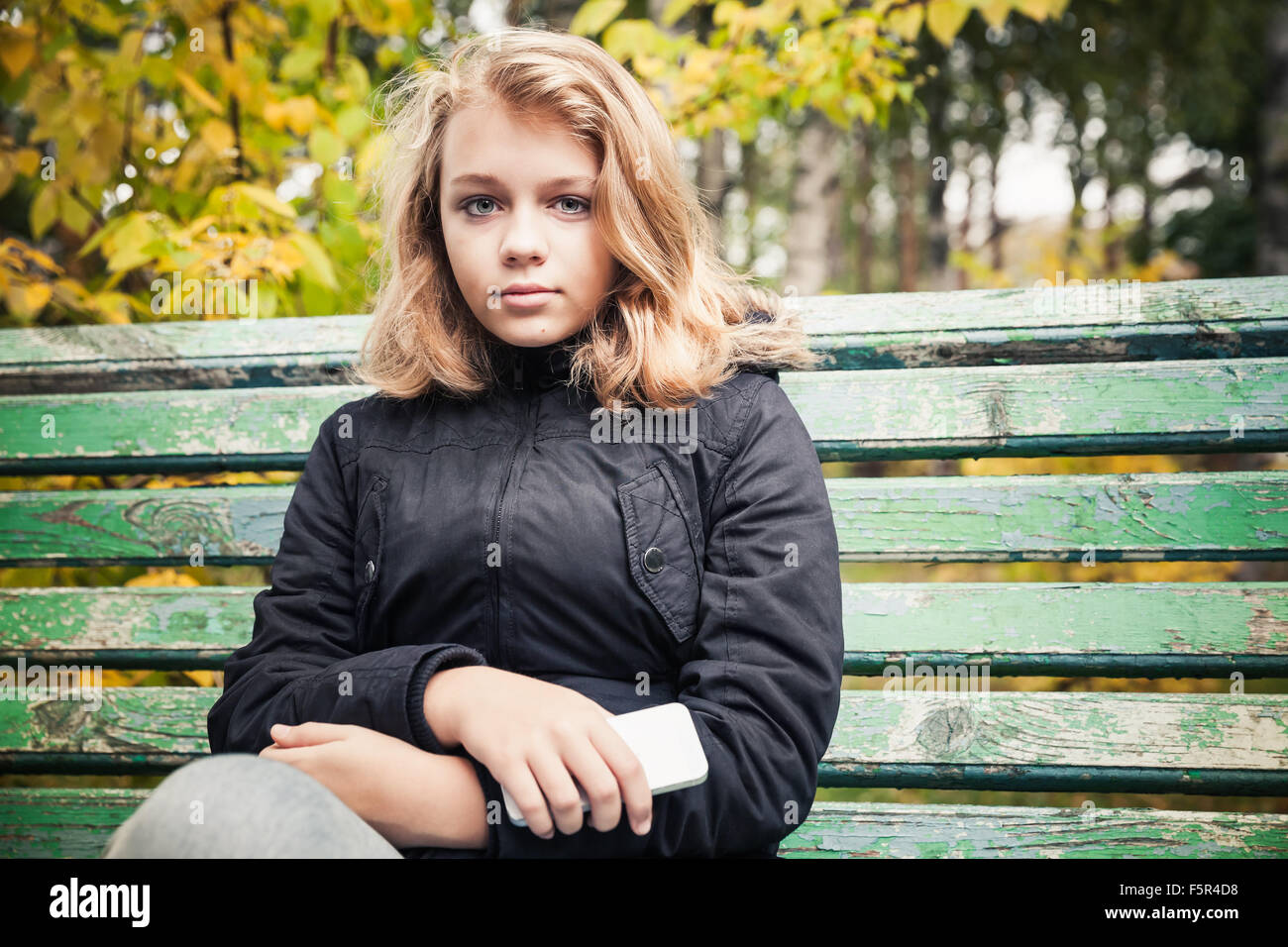 Bella bionda caucasica ragazza adolescente in giacca nera seduta sul parco verde di banco con smartphone nelle mani Foto Stock