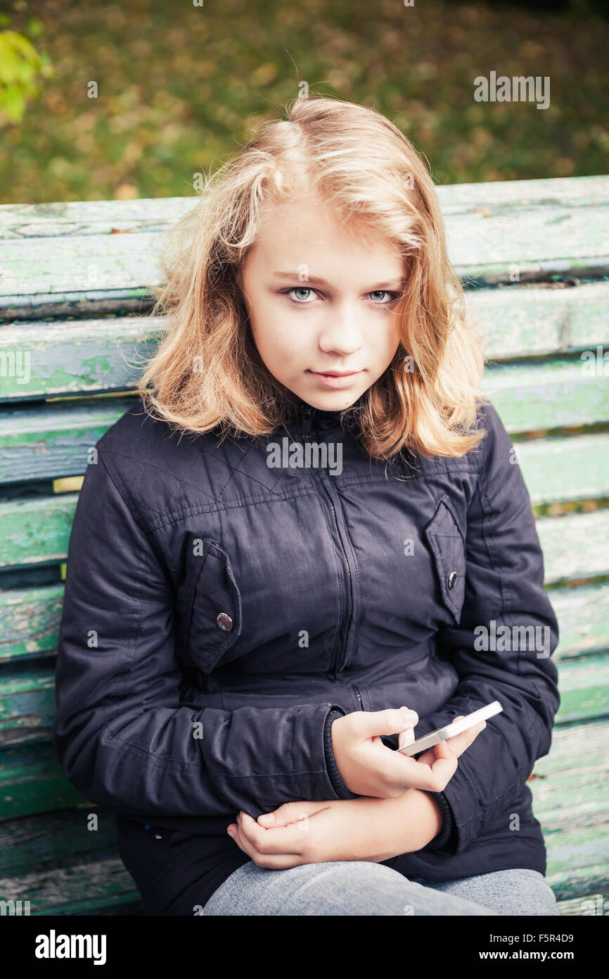 Bella bionda caucasica ragazza adolescente in giacca nera seduta sul vecchio parco verde di banco con smartphone nelle mani Foto Stock