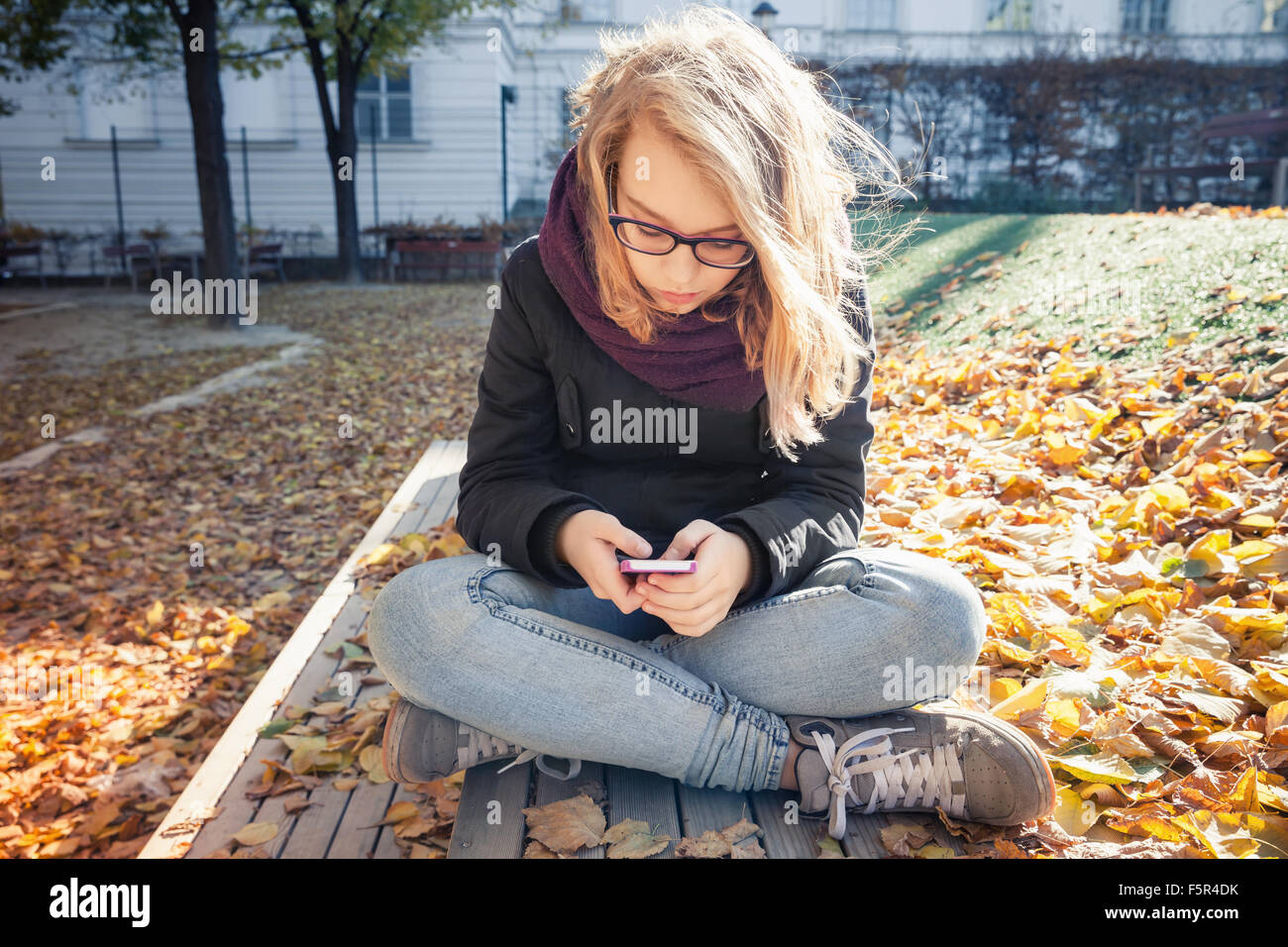 Carino bionda caucasica ragazza adolescente in jeans e giacca nera seduto su una panchina nel parco e utilizza lo smartphone, outdoor autunno ritratto Foto Stock