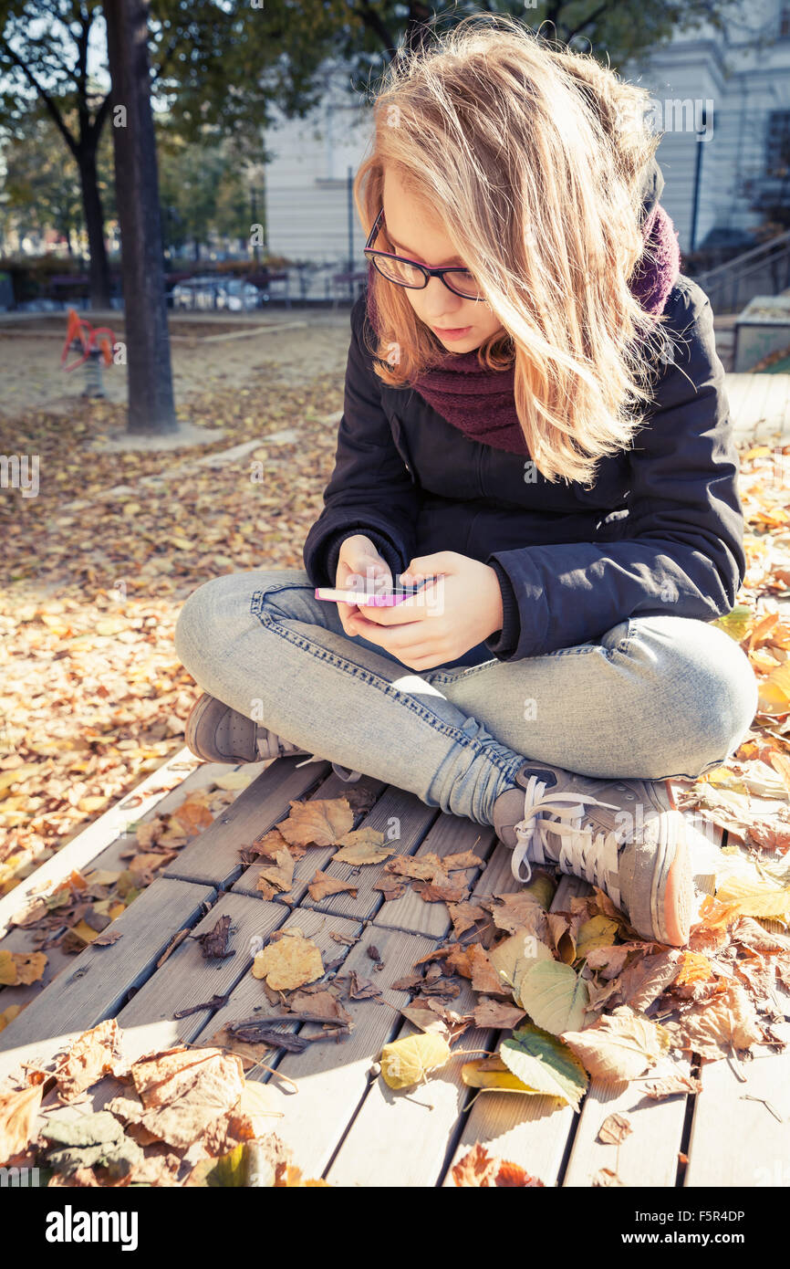 Carino bionda caucasica ragazza adolescente in jeans e giacca nera seduta in legno su una panchina nel parco e utilizza lo smartphone, outdoor autunno po Foto Stock