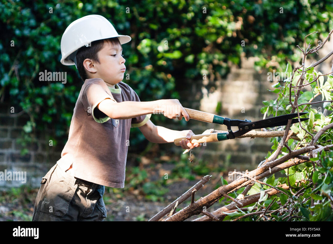 Bambino caucasico, boy, indossando di sicurezza bianca cappello e t-shirt, cercando di tagliare rami con grande giardino cesoie. Espressione seria. Foto Stock