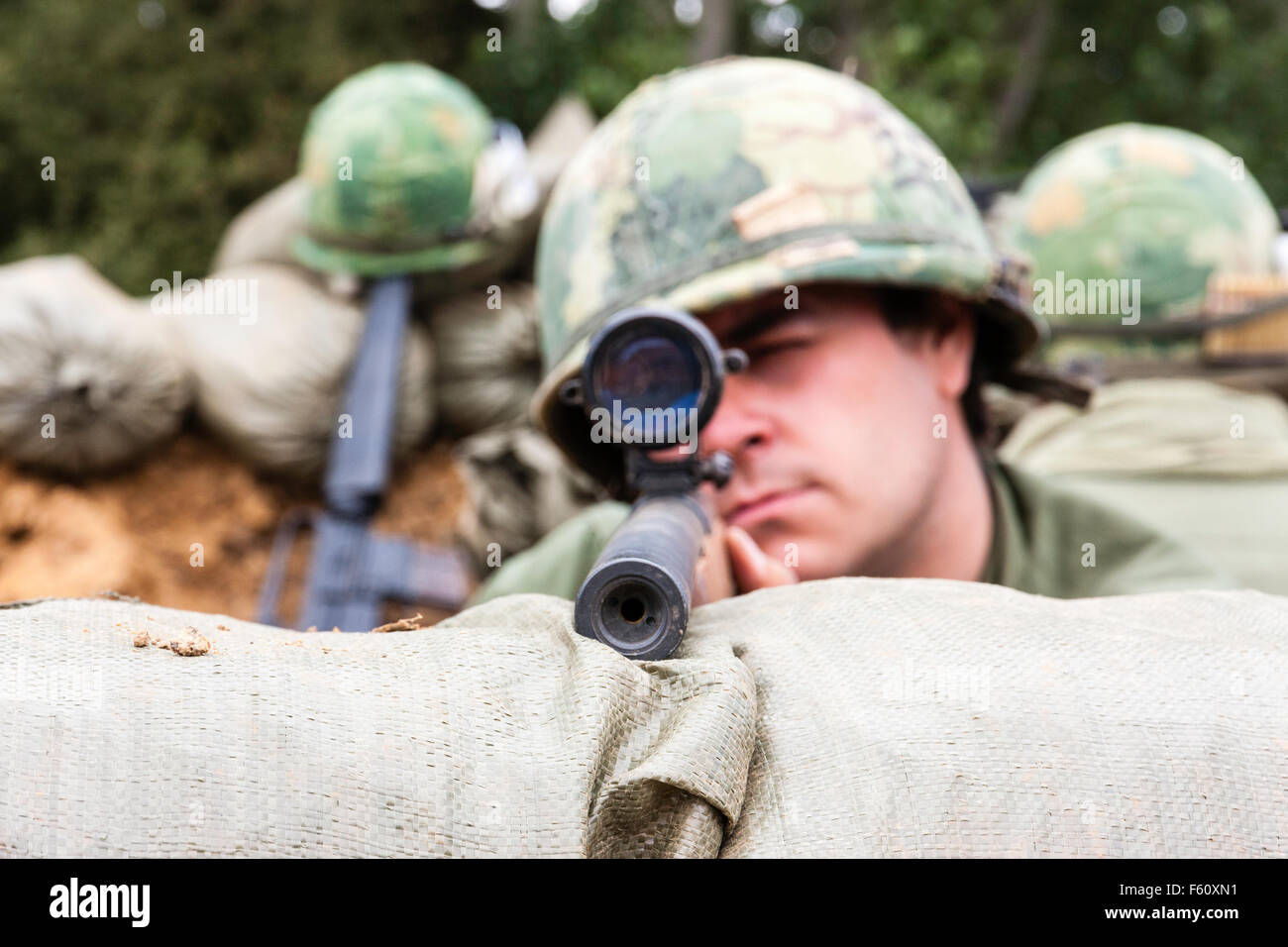 Guerra del Vietnam rievocazione gruppo. Soldato americano, cecchino testa sopra dei sacchi di sabbia, guardando il telespettatore attraverso la portata tenendo obiettivo. Close up. Contatto visivo. Foto Stock