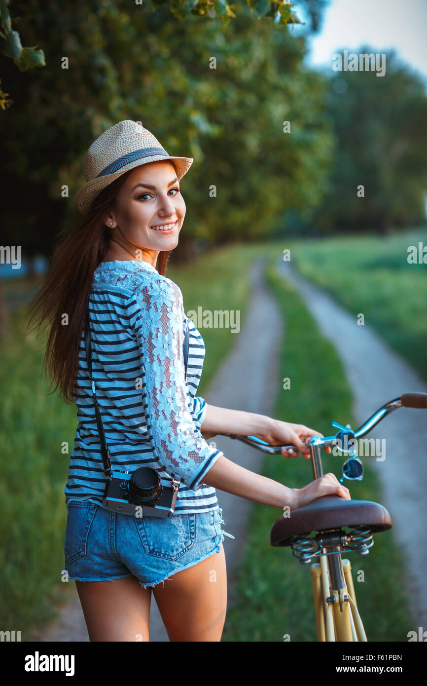 Bella giovane donna in un cappello con una bicicletta in un parco. All'aperto Foto Stock