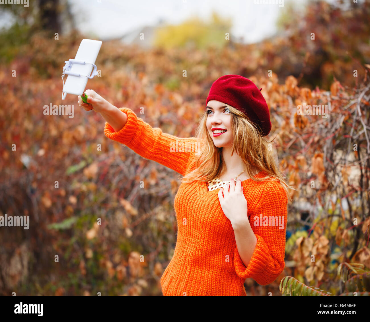 Hipster bella ragazza in un berretto e un maglione in autunno rende il parco auto ritratto su un telefono cellulare. Selfie. Ragazza detiene selfie stick. Foto Stock