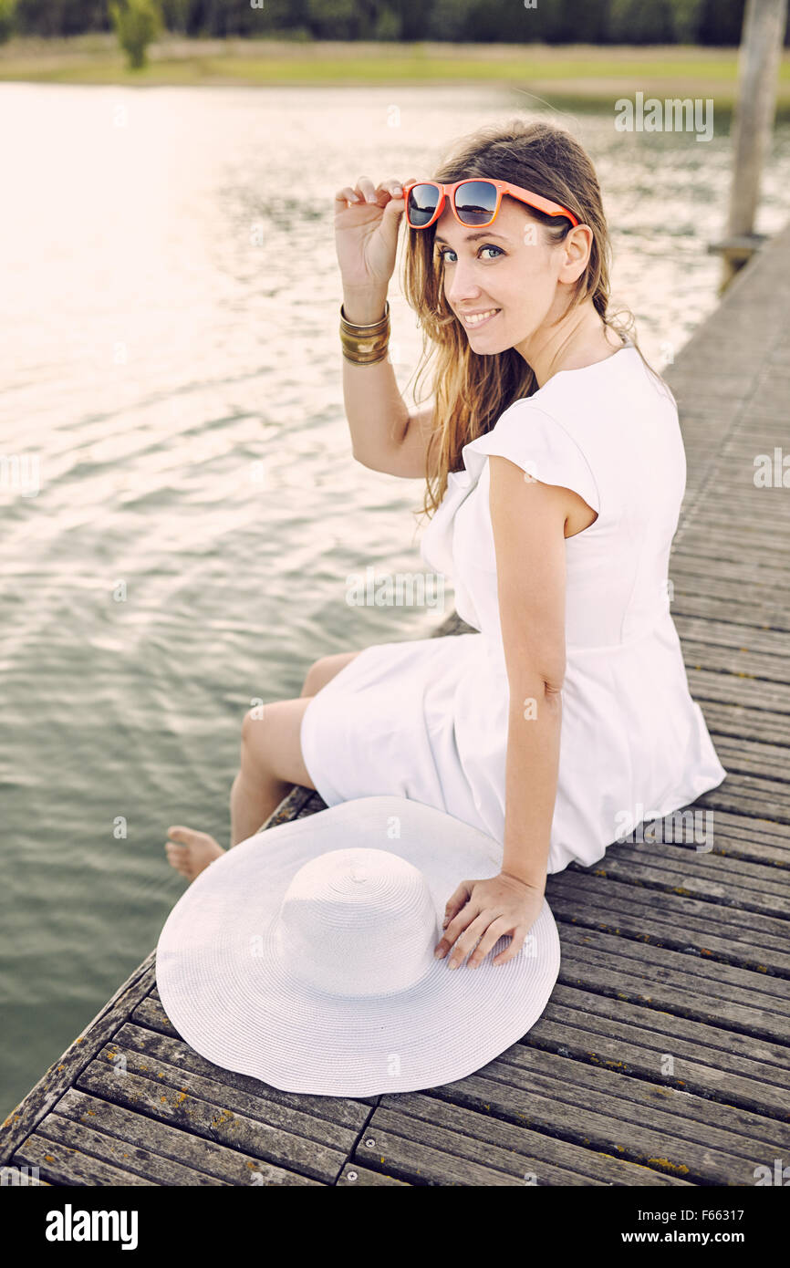 Ritratto di una donna seduta su un dock con abito bianco e hat e vetro arancione Foto Stock