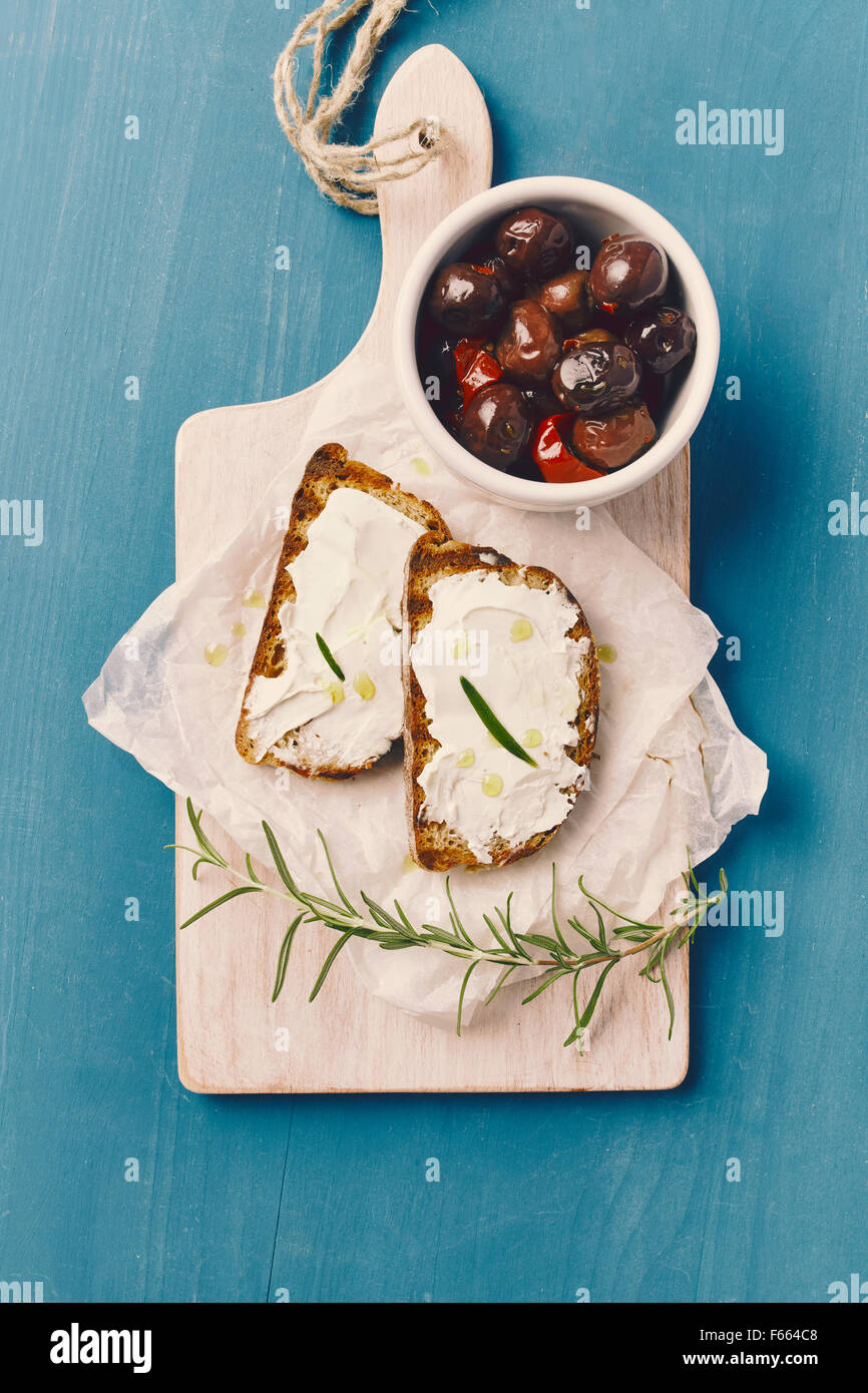 Vista dall'alto di due fette di pane grigliato con formaggio spalmabile e olio su un bianco tagliere, su un blu tavolo in legno con ol Foto Stock