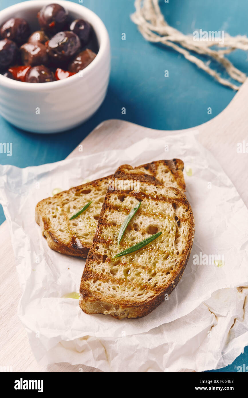 Due fette di pane grigliato con olio su un bianco tagliere, sul blu di un tavolo di legno con le olive in un recipiente bianco un rosmarino Foto Stock