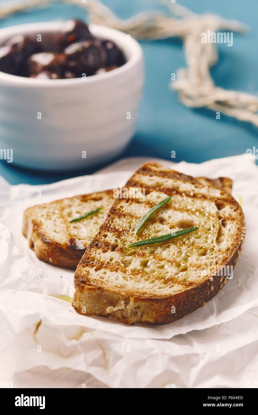 Due fette di pane grigliato con olio su un bianco tagliere, sul blu di un tavolo di legno con le olive in un recipiente bianco un rosmarino Foto Stock
