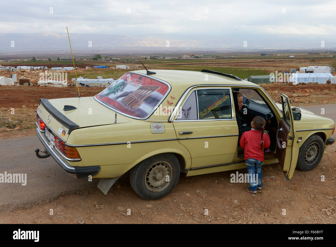 Il Libano Beqaa valley, Deir el Ahmad, campo per rifugiati siriano, antico tedesco Mercedes Benz 200 D auto con antico tedesco codice paese Kennzeichen D , bandiera libanese e l'immagine del tempio di Baalbek, sfondo Anti montagne del Libano e Siria Foto Stock