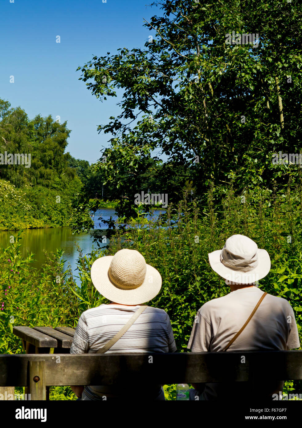 Coppia di anziani che indossa cappello da sole seduto su una panca in legno godendo di una calda giornata estiva in prossimità di un lago Foto Stock