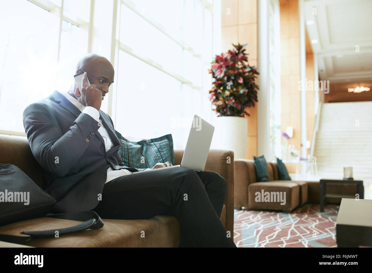 Imprenditore seduti sul divano a lavorare utilizzando il telefono cellulare e il computer portatile. African executive maschio in attesa nella hall dell'hotel. Foto Stock