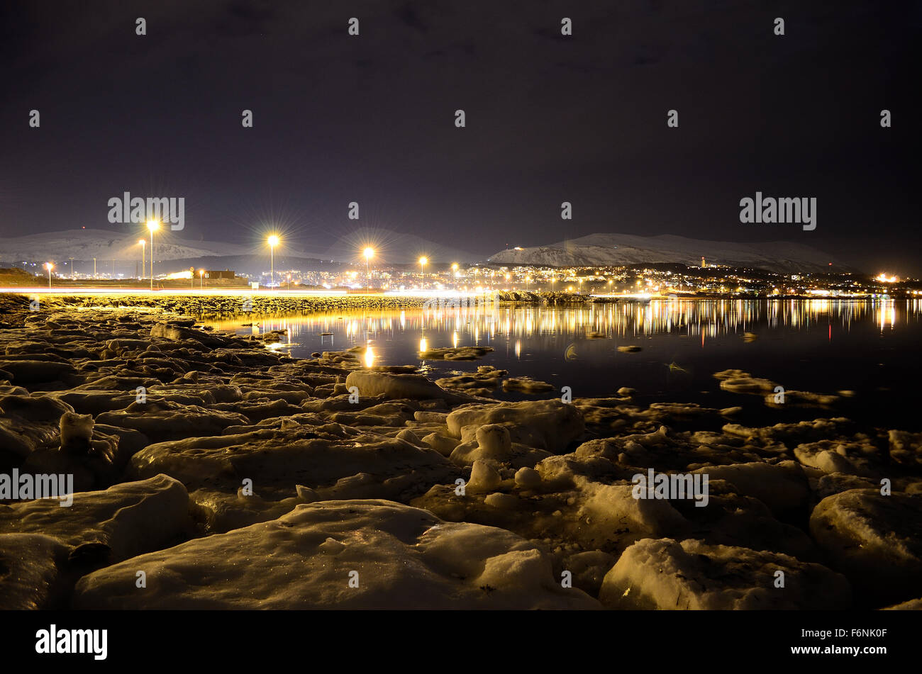 Lungo litorale innevato con luce riflettendo sulla superficie con tromsoe città illuminazione notturna in background Foto Stock