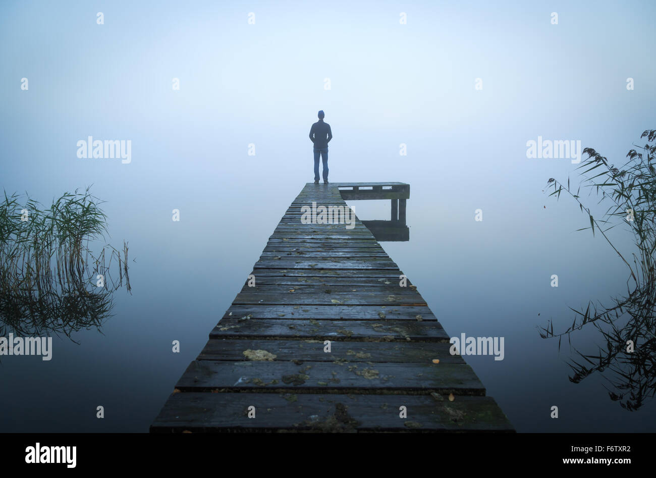 Uomo in piedi su un molo a un lago durante una nebbia, grigia mattina. Foto Stock