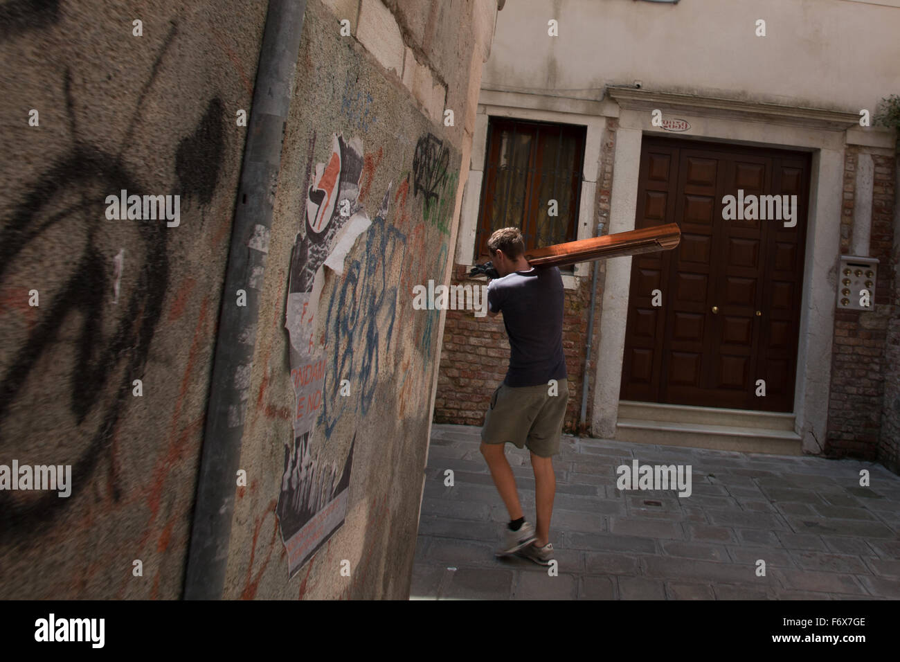 Uomo che trasporta un lungo bastone di legno davanti a un muro ricoperto di graffiti in una stradina di Venezia, Veneto, Italia Foto Stock