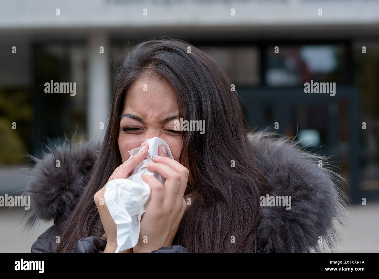 Donna con un inverno stagionali raffreddore e influenza in piedi all'aperto su una strada urbana in un mantello peloso soffia il naso su un handkerch Foto Stock