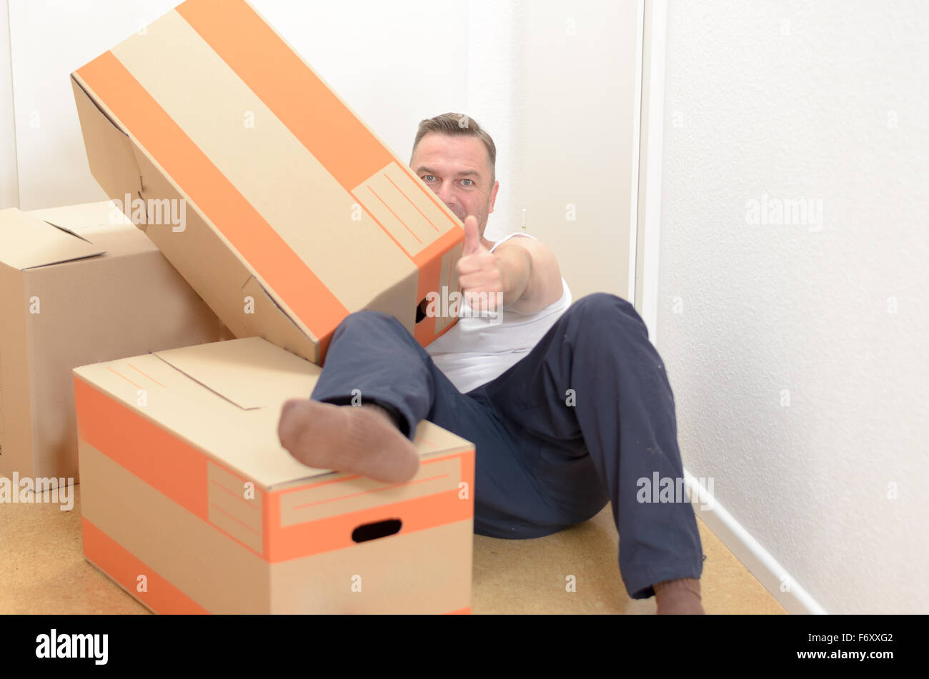 Uomo di casa in movimento rilassante tra una collezione di scatole di cartone compresso con i suoi beni sdraiato sul pavimento dando un pollice in u Foto Stock