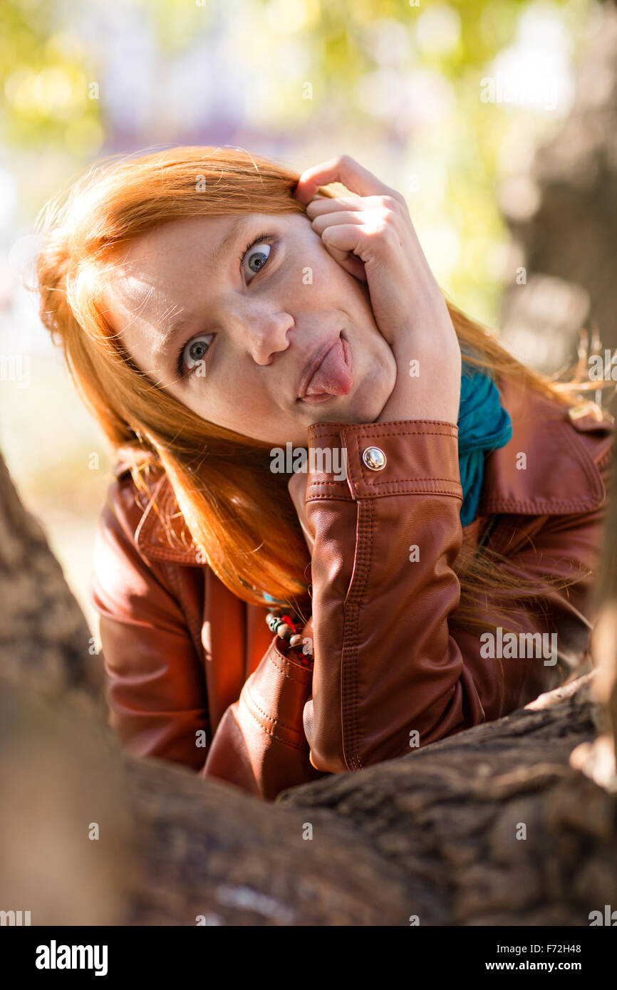Abbastanza divertente redhead girl rendere divertente la faccia e che mostra la linguetta che pongono nel parco vicino tree Foto Stock