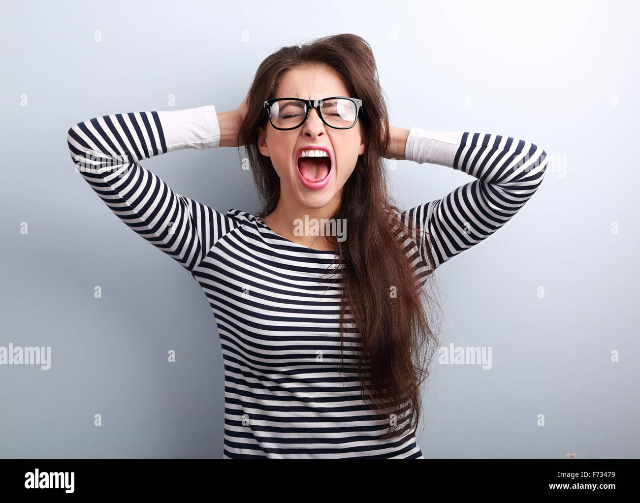 Rabbia giovane donna di affari in bicchieri forte urlando selvaggio con bocca aperta e tenendo la testa le mani su sfondo blu Foto Stock