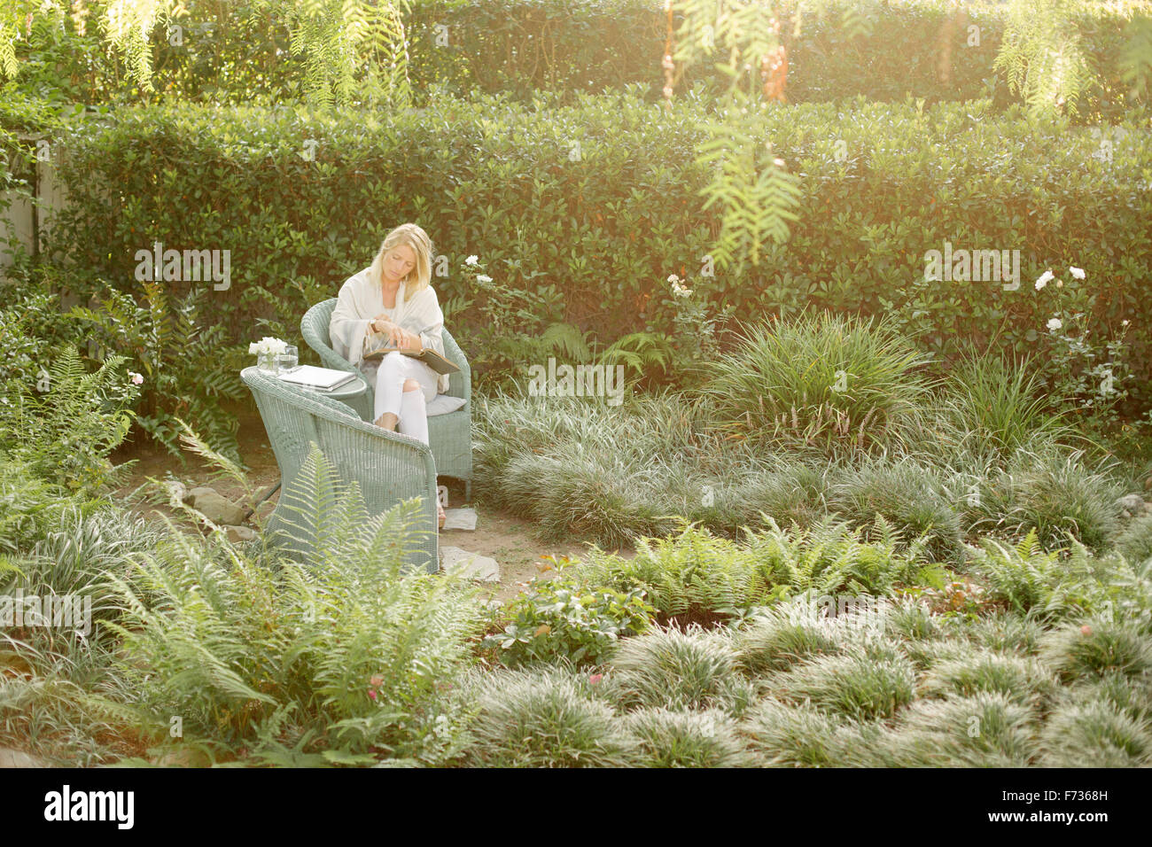 Donna bionda seduto in una sedia di vimini in un giardino, la lettura. Foto Stock